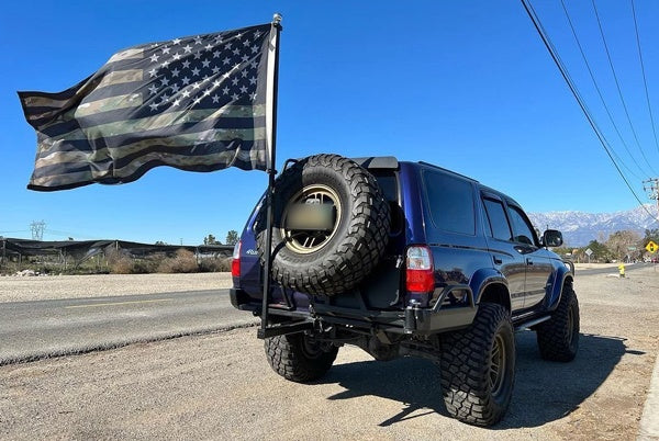 Blue SUV with a rear spare tire carrier flying a full-size camo American flag from a FanPole on the extended dummy hitch, with mountains behind
