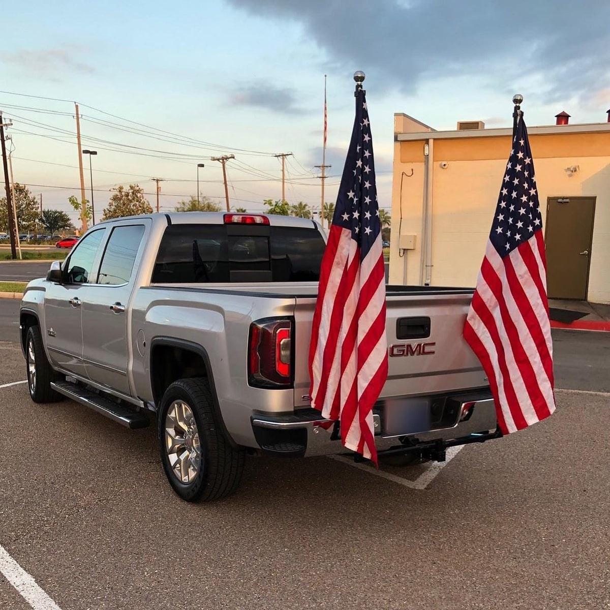 Silver pickup truck in a parking lot with two FanPole flag poles mounted at the rear hitch, each flying a full-size American flag.