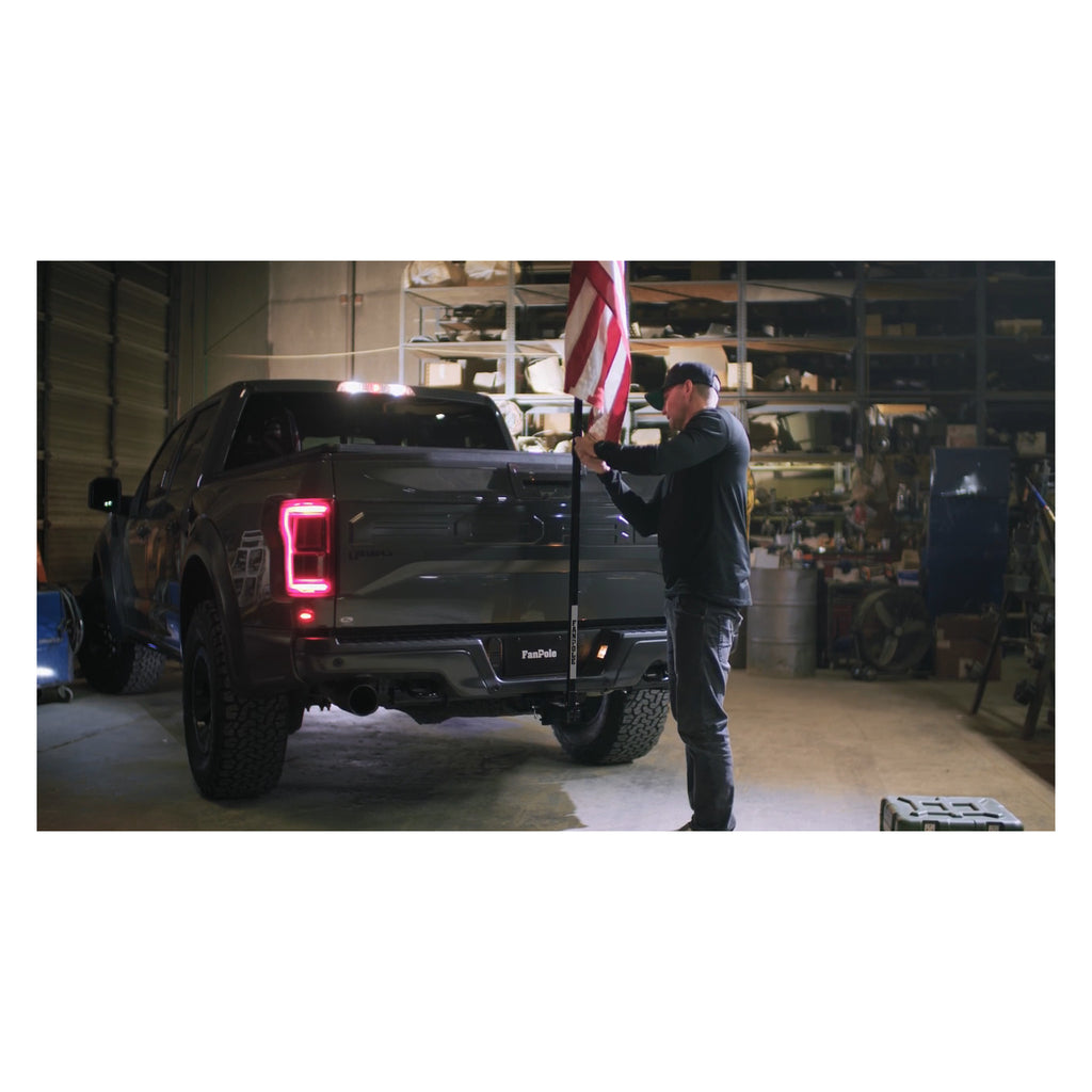 Man setting up a FanPole flag pole in the rear hitch of a dark pickup truck inside a workshop, with a full-size American flag on the pole.
