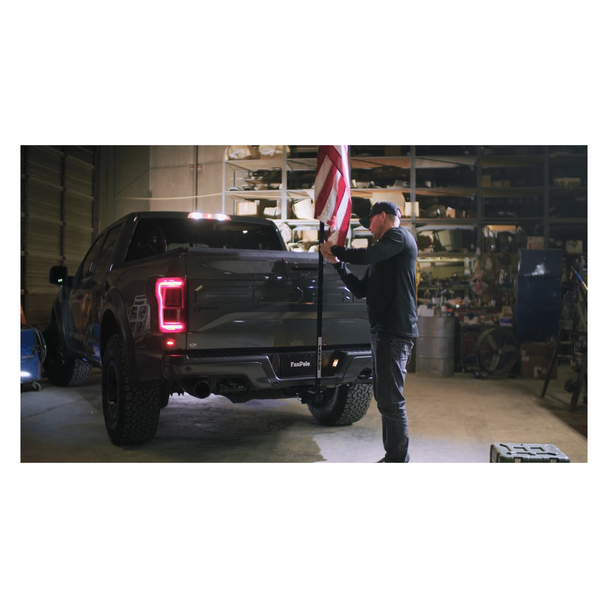 Man setting up a FanPole flag pole in the rear hitch of a dark pickup truck inside a workshop, with a full-size American flag on the pole.